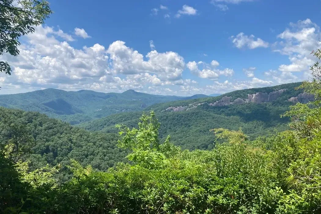 Wide ridgeline view from the upper section of Bennett Gap Trail in Pisgah National Forest, looking over rolling green mountains and exposed rock outcrops under a partly cloudy sky
