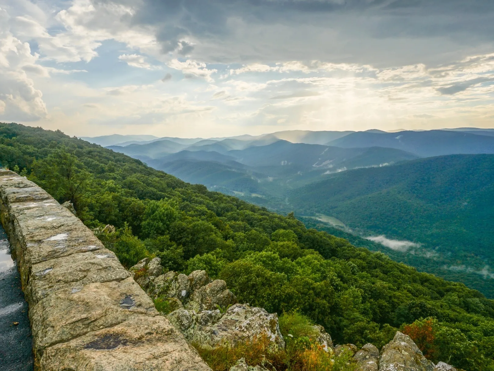 View from a stone-walled Blue Ridge Parkway overlook across layered forested ridgelines under a cloud-streaked sky