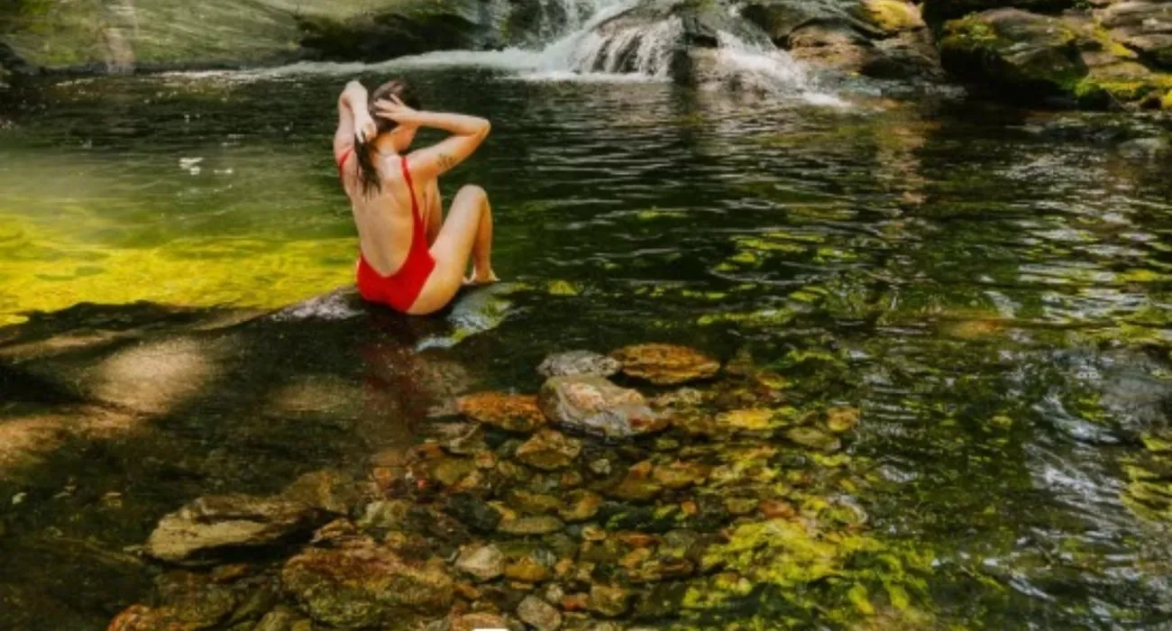 A visitor in a red swimsuit cooling off in a clear green creek pool below a small cascade, one of the swimming spots along the trail to Daniel Ridge Falls
