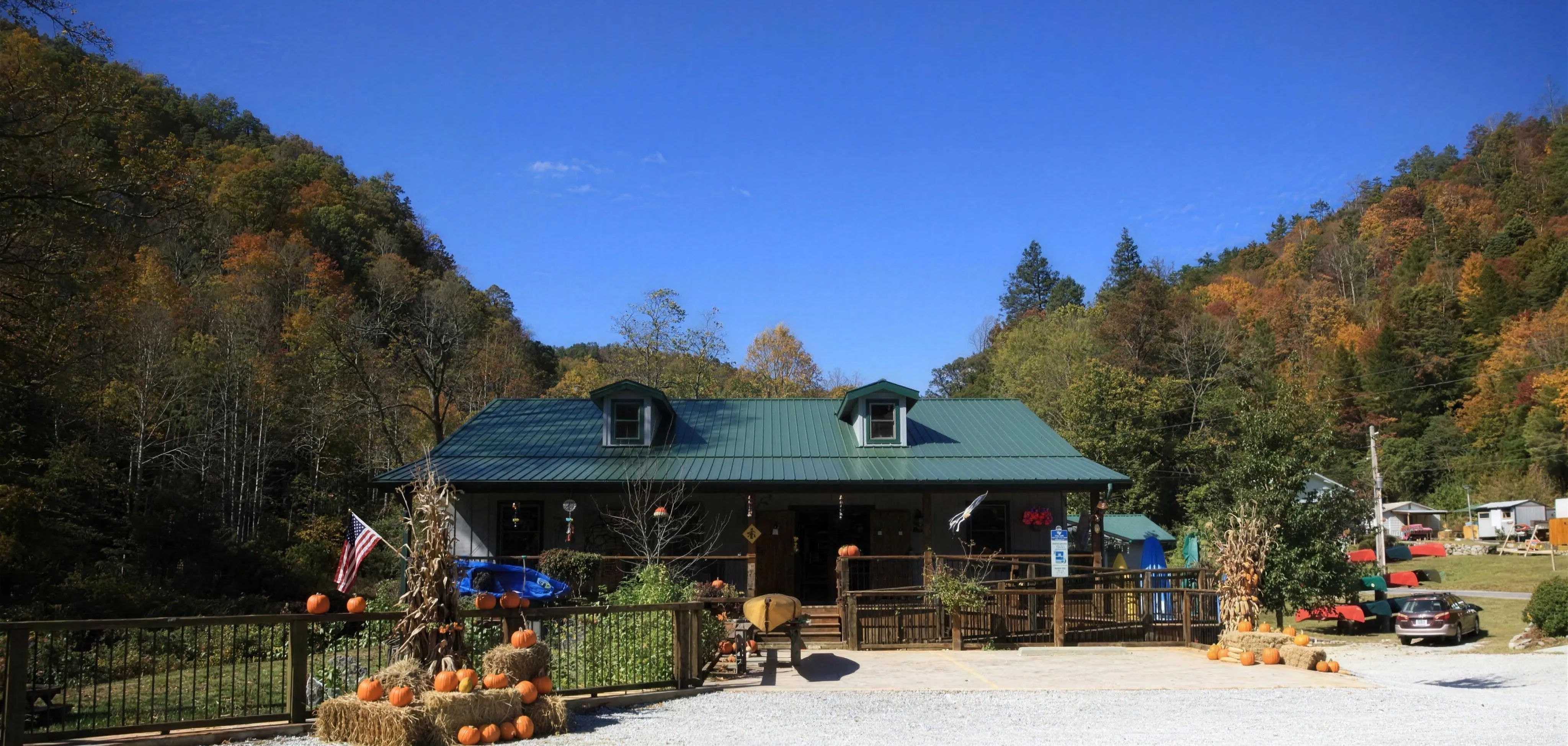 The Headwaters Outfitters shop at the Forks of the French Broad in Rosman, NC, on a clear blue-sky autumn day, with corn stalks, pumpkins, and hay bales out front and a fleet of canoes and kayaks visible to the right