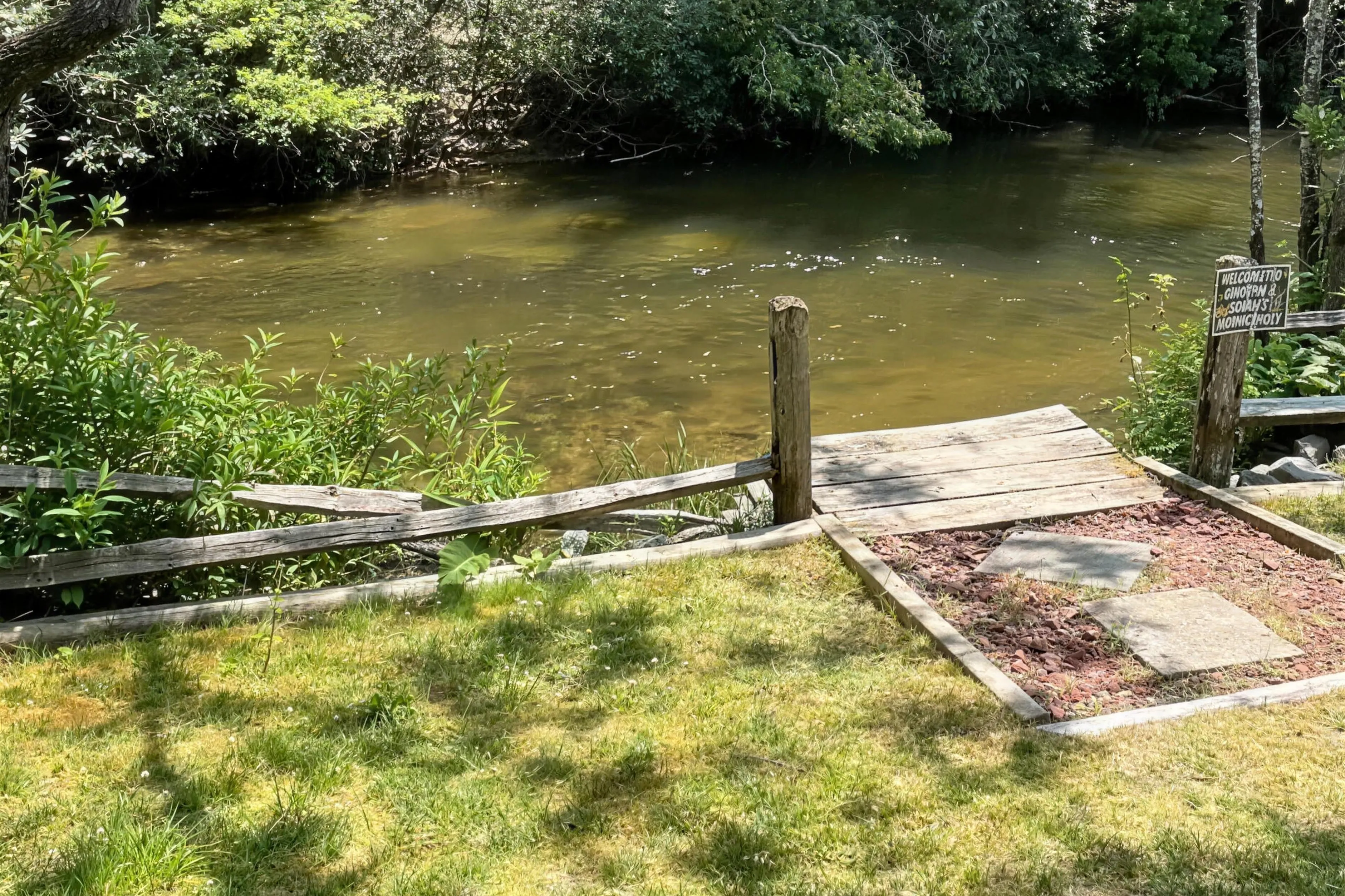 A riverside campsite at Headwaters Outfitters on the upper French Broad River, with a wooden dock, split-rail fence, and a welcome sign for the campground fishing hole