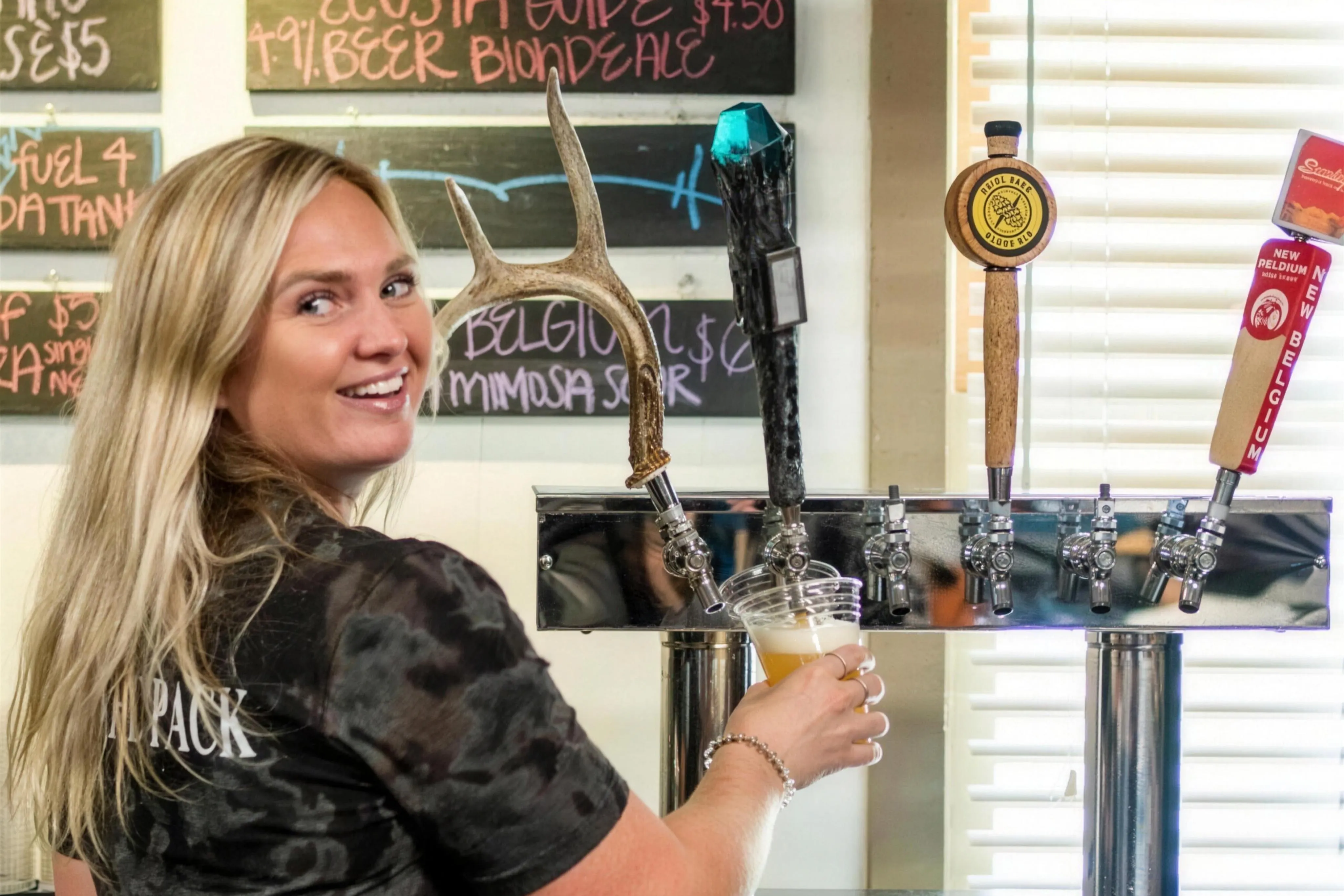 A smiling bartender pulling a pint at the Forks of the River Taproom at Headwaters Outfitters, with antler tap handles, chalkboard beer menus, and New Belgium Brewing taps visible behind her