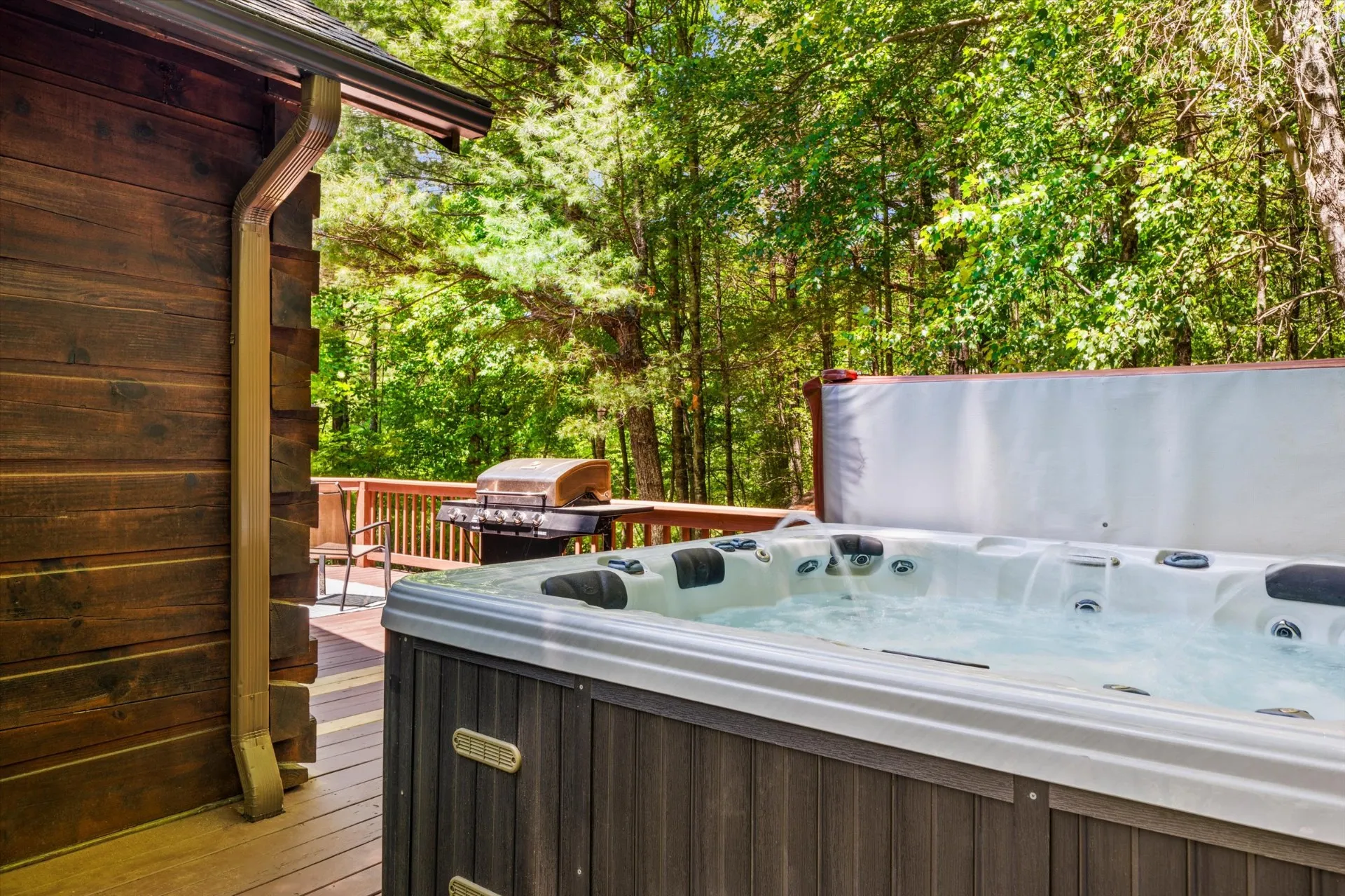 Covered hot tub on the wood deck beside the cabin, with a gas grill and forest views
