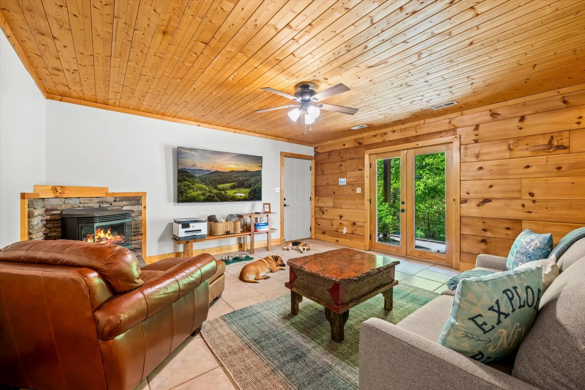 Lower level family room with stone gas fireplace, leather sofa, wood-paneled walls and ceiling, mounted TV, and french doors opening to the woods