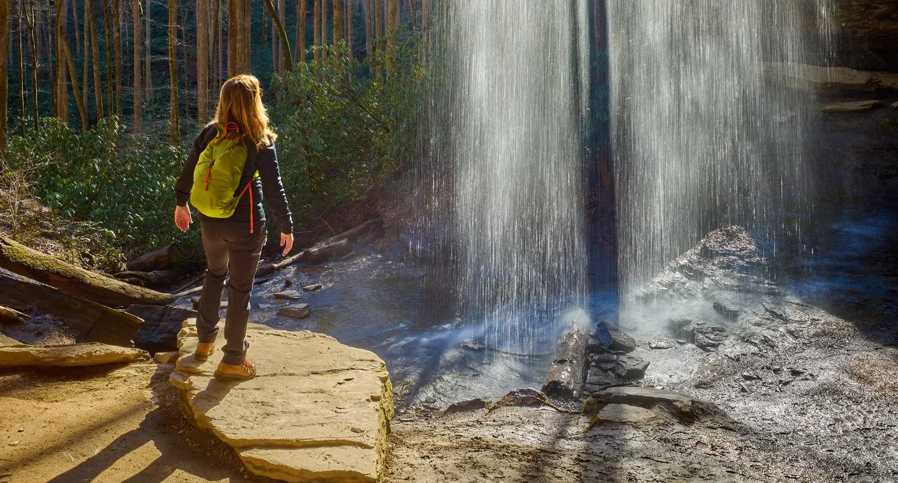 A hiker in a yellow pack standing on the rock ledge at the base of Moore Cove Falls, looking up at the veil pouring over the overhang