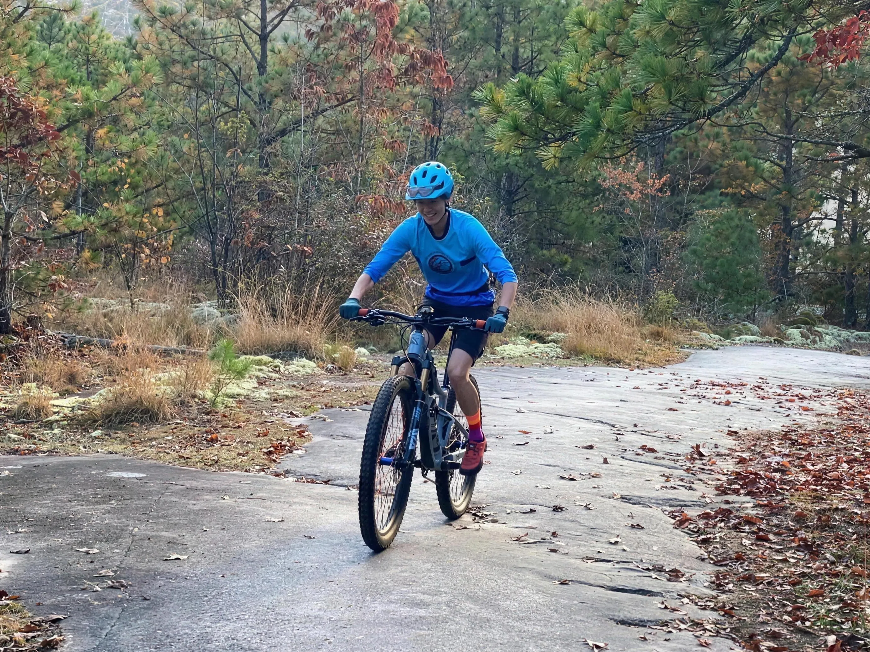 A smiling mountain biker in a blue helmet and jersey riding across an open granite slab with fall leaves