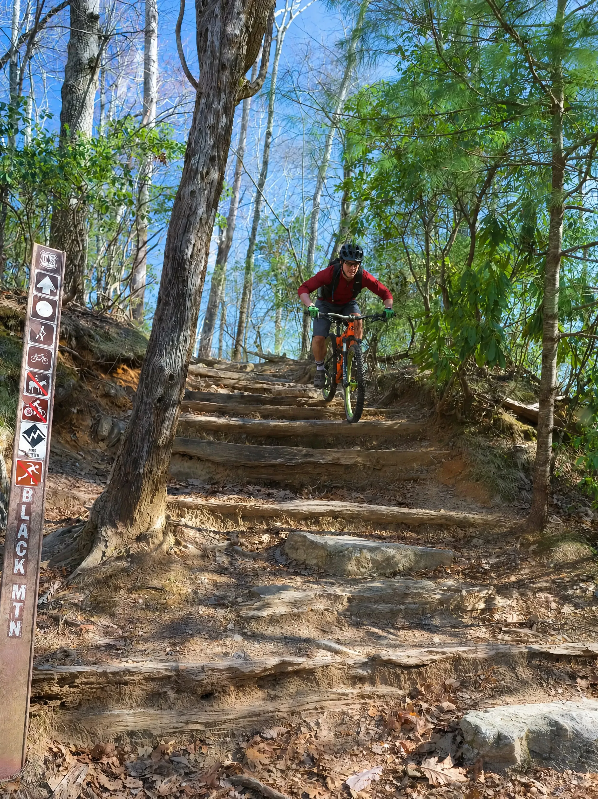 A mountain biker in a red jersey riding down steep log steps past a Black Mtn trail post