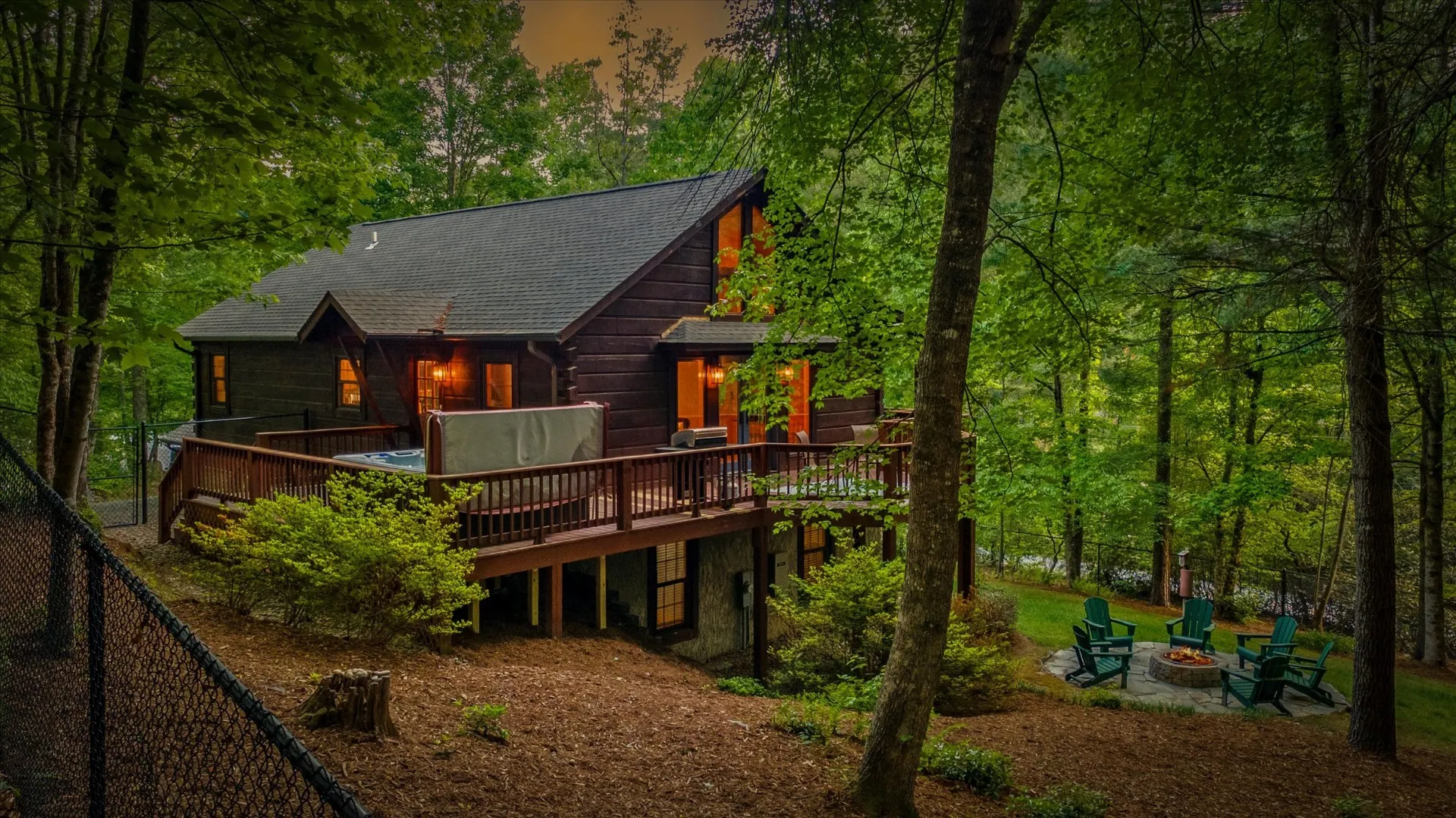 Pisgah Base Camp cabin at dusk — log exterior with lit windows, wraparound deck with hot tub, and stone fire pit ringed by Adirondack chairs in the Pisgah Forest